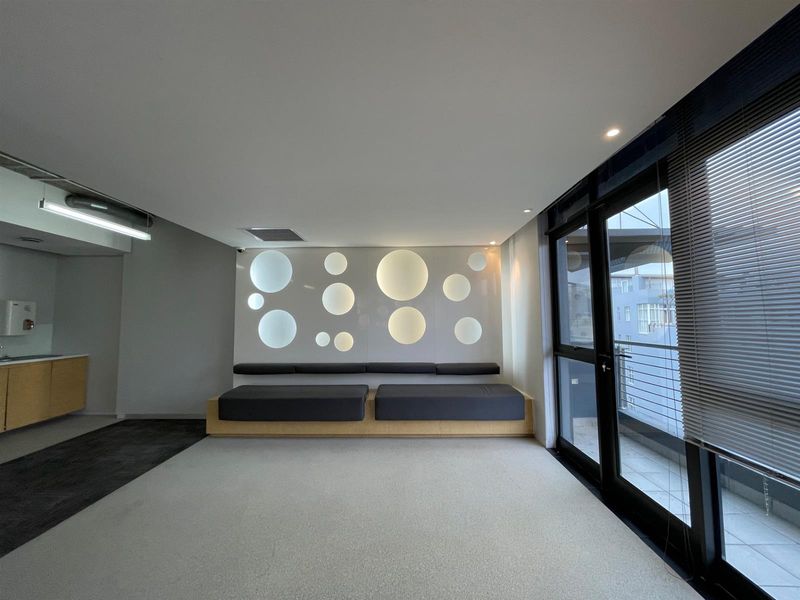 Modern office space featuring built-in bench seating with decorative circular wall lighting, a small kitchenette area, and large glass doors leading to a balcony.