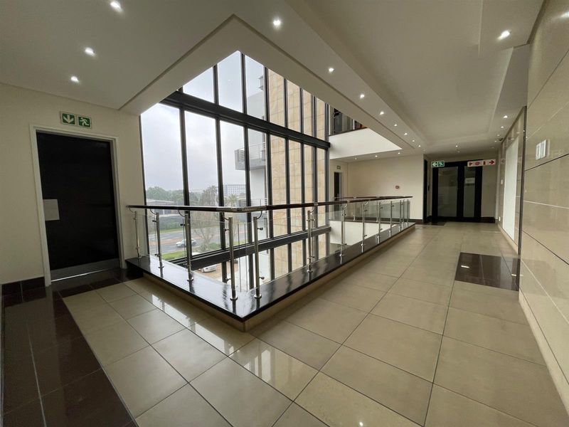 Modern office building hallway featuring a double-volume glass curtain wall, polished tile flooring, and a contemporary stainless steel and glass balustrade.