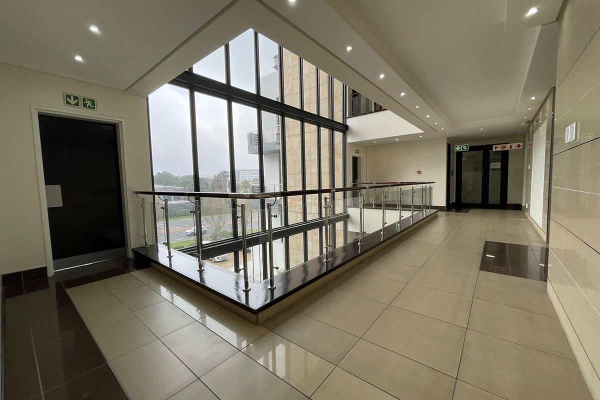 Modern office building hallway featuring a double-volume glass curtain wall, polished tile flooring, and a contemporary stainless steel and glass balustrade.