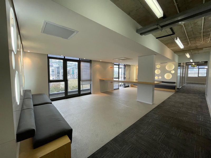 Modern office space featuring a mix of carpet and vinyl flooring, exposed concrete ceilings, built-in bench seating, and large glass doors providing natural light.