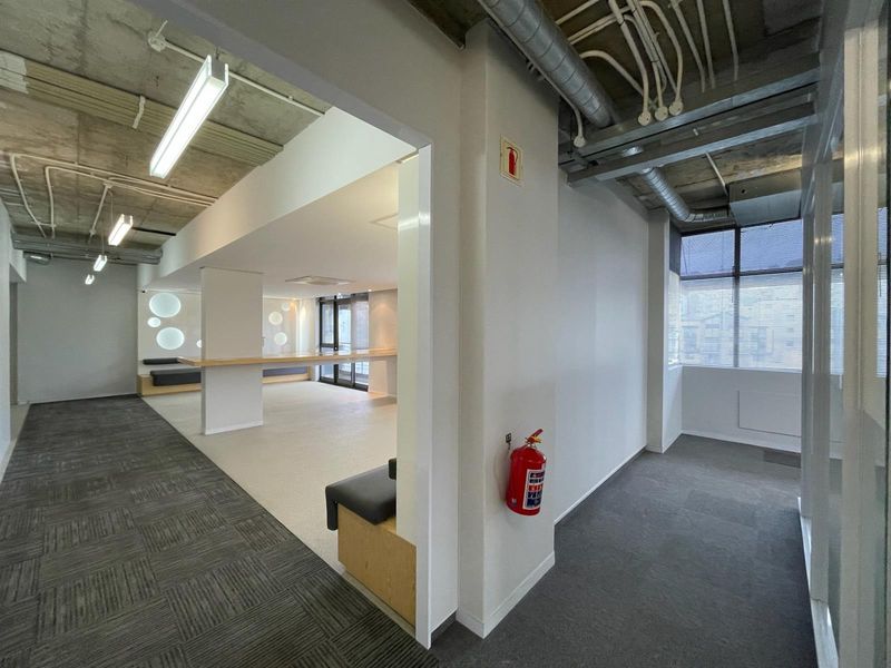 Modern open-plan office space featuring exposed industrial ceilings, built-in counter and bench seating, carpeted flooring, and large windows providing ample natural light.