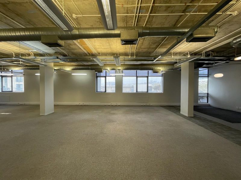 A spacious, open-plan office area featuring an industrial-style exposed concrete ceiling with visible ducting, large windows, and neutral carpeting.