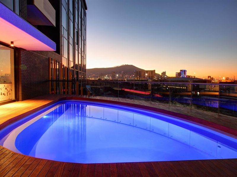 A modern elevated swimming pool with timber decking and glass balustrades, offering stunning city and mountain views at dusk.