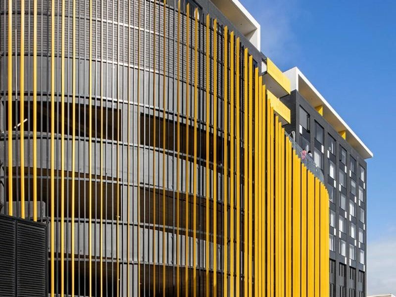 Modern apartment building exterior featuring a striking yellow and grey facade with vertical louvres and integrated parking structure.