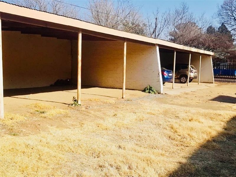 An open-sided carport structure with timber support posts and a corrugated metal roof, providing covered parking space on an unpaved lot.