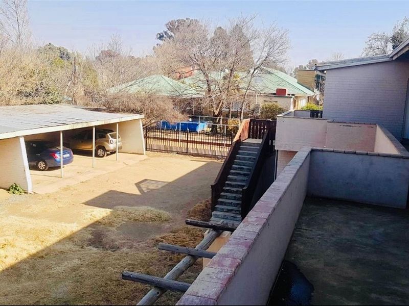 Rear exterior view showing a carport, elevated balcony with staircase access, and a large open yard area with boundary walling.