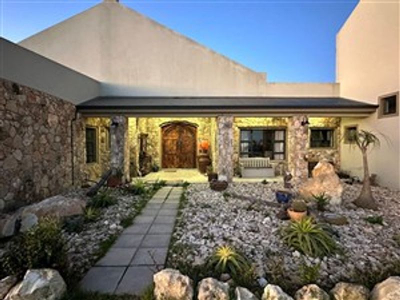Front entrance of a farm-style property featuring stone cladding, a paved walkway, and a prominent timber front door set within a landscaped rock garden.