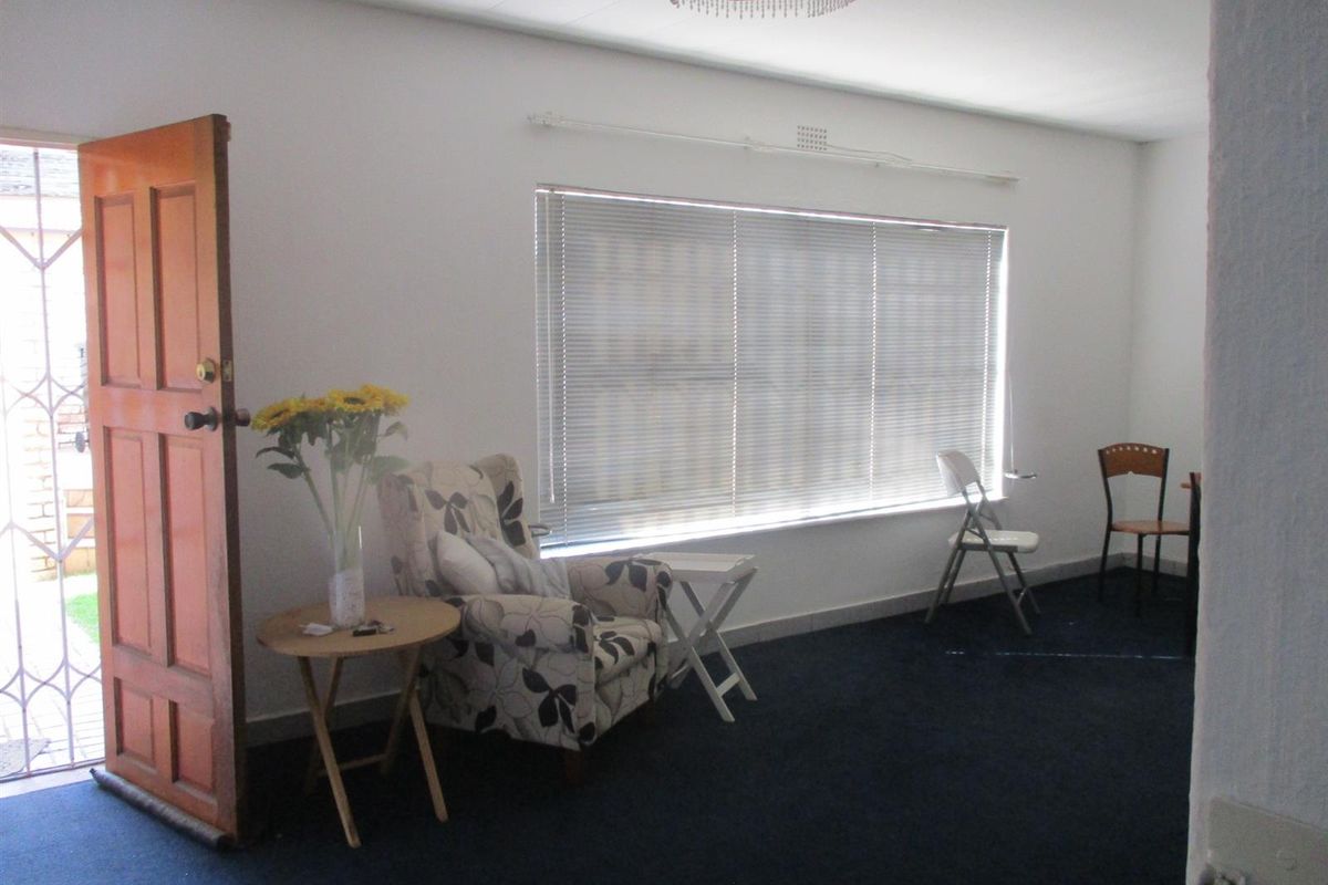 A living room featuring dark carpeting, a large window with venetian blinds, and a timber entrance door with an integrated security gate.