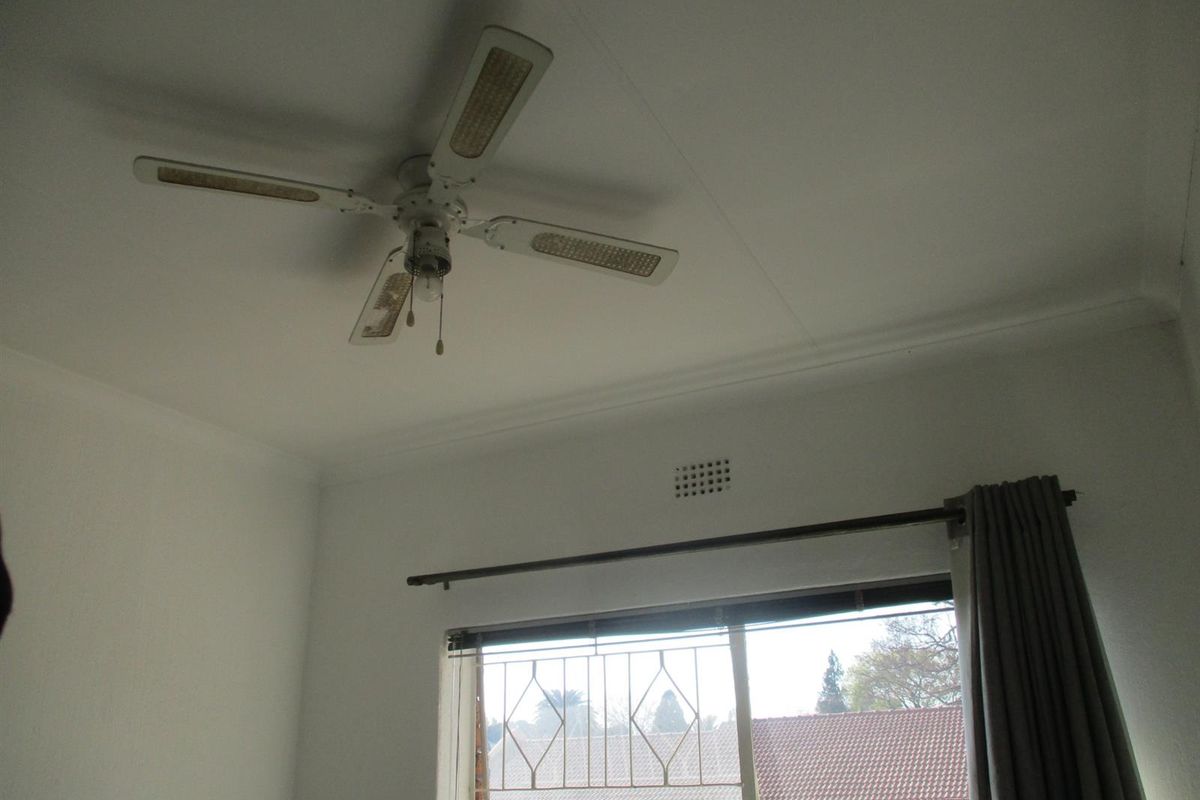 A bedroom interior featuring a ceiling fan, security gate on the window, and curtain rod, showing signs of age and wear.
