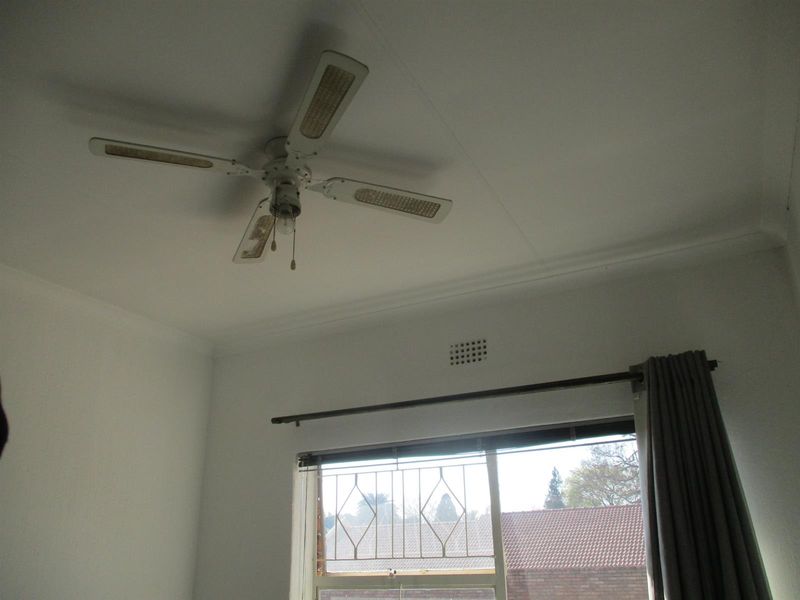 A bedroom interior featuring a ceiling fan, security gate on the window, and curtain rod, showing signs of age and wear.