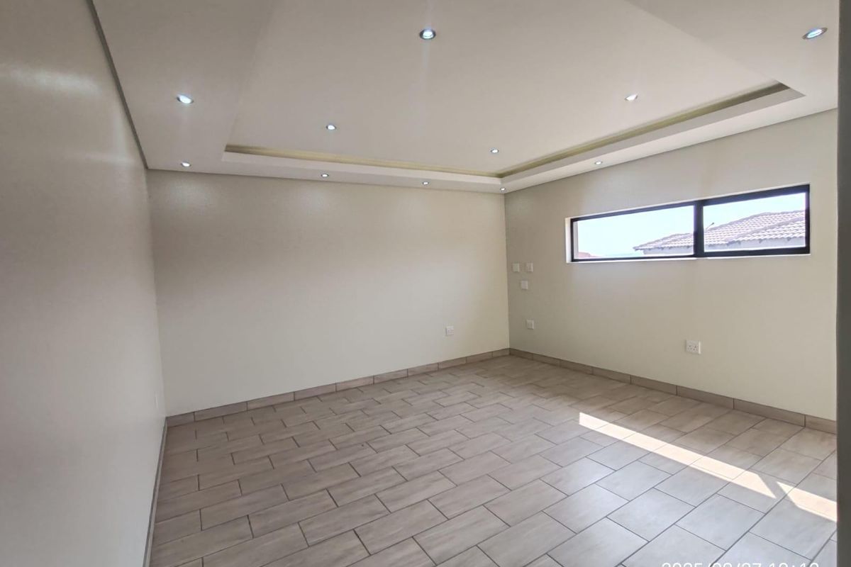 A bright, empty living room featuring modern recessed ceiling lighting, neutral tiled flooring, and a horizontal window providing natural light.
