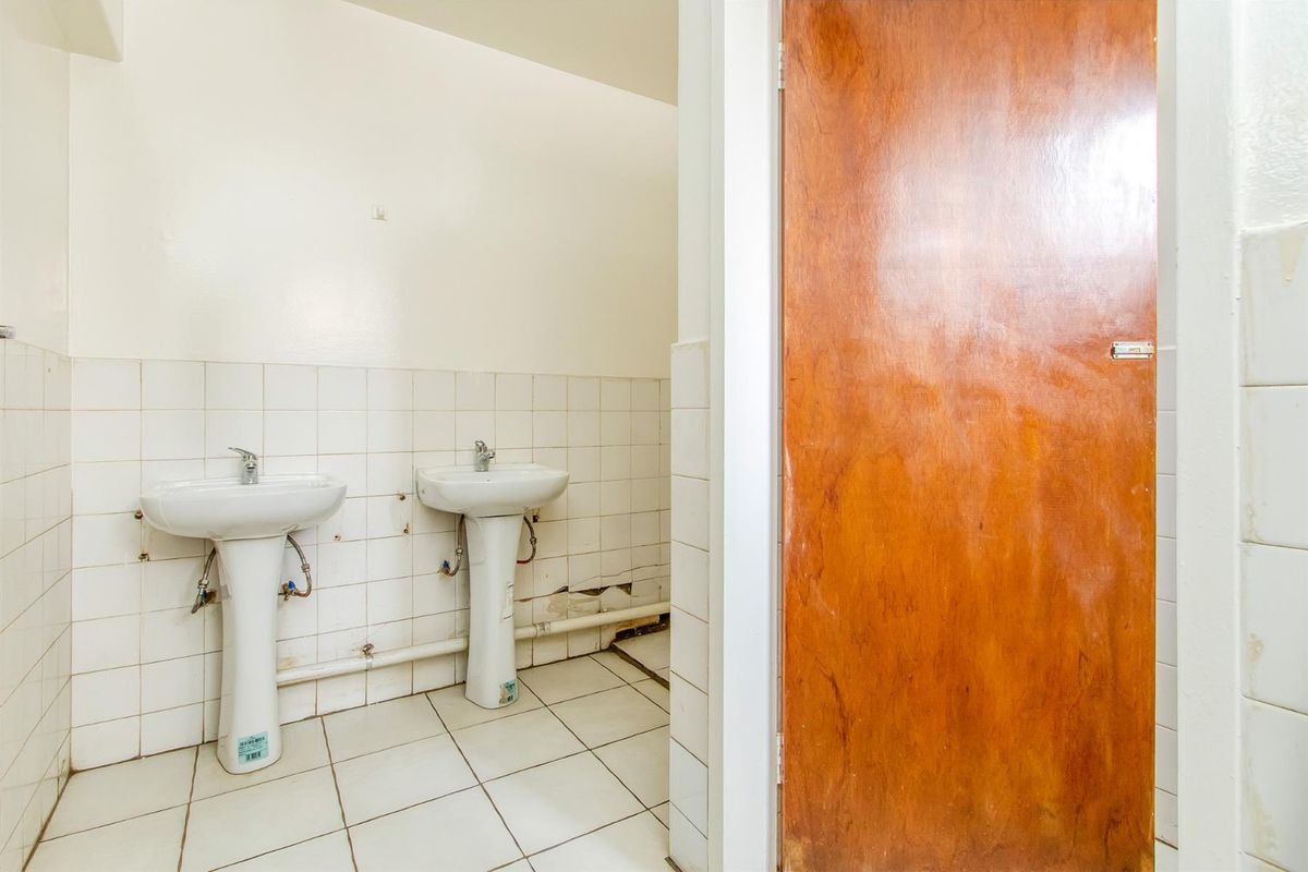 A commercial bathroom facility featuring dual pedestal sinks with tiled splashbacks and a wooden door, currently showing signs of wear and maintenance requirements.