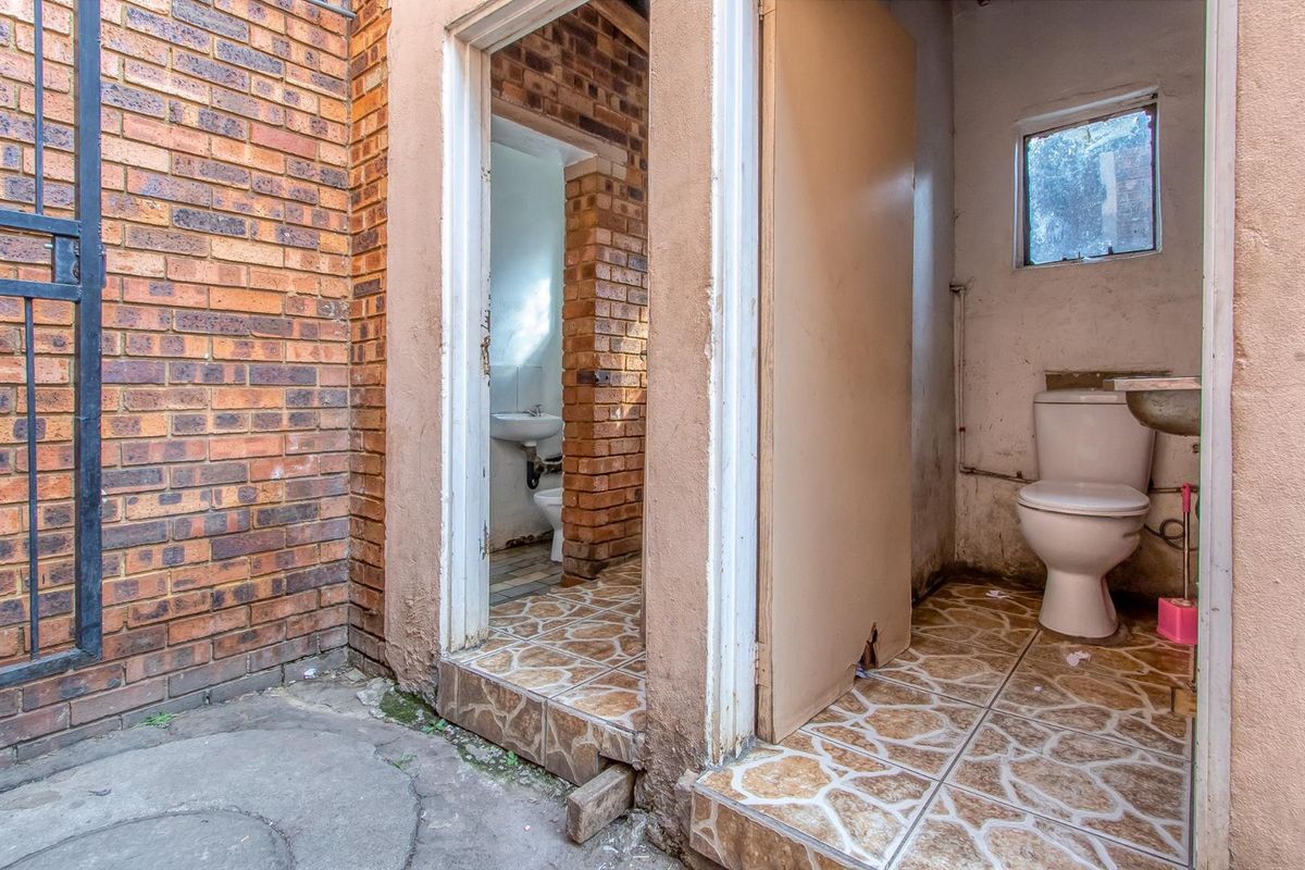 Two small, separate toilet cubicles with tiled flooring and basic sanitary fixtures, accessible from an exterior brick-walled area.