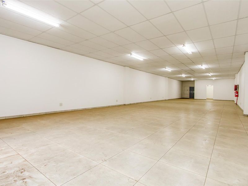 A spacious, empty commercial open-plan area featuring tiled flooring, a suspended ceiling with fluorescent lighting, and a wall-mounted fire extinguisher.