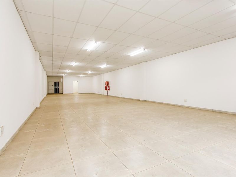 A spacious, empty commercial open-plan area featuring ceramic tiled flooring, a suspended ceiling with fluorescent lighting, and a wall-mounted fire extinguisher.