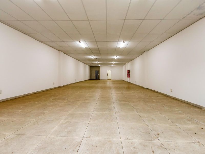 A large, empty commercial open-plan space featuring tiled flooring, a suspended ceiling with integrated lighting, and a wall-mounted fire extinguisher.