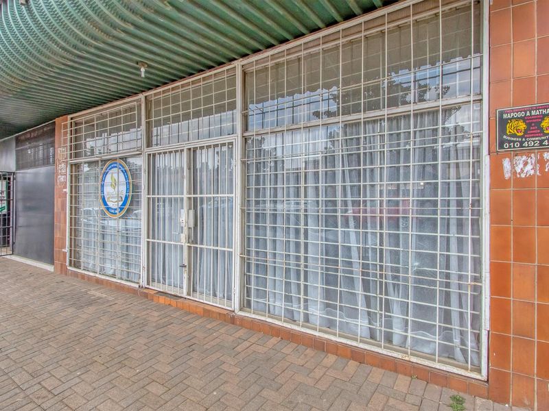 Commercial storefront featuring a tiled facade, large glass windows with security bars, and a corrugated metal awning over a paved walkway.