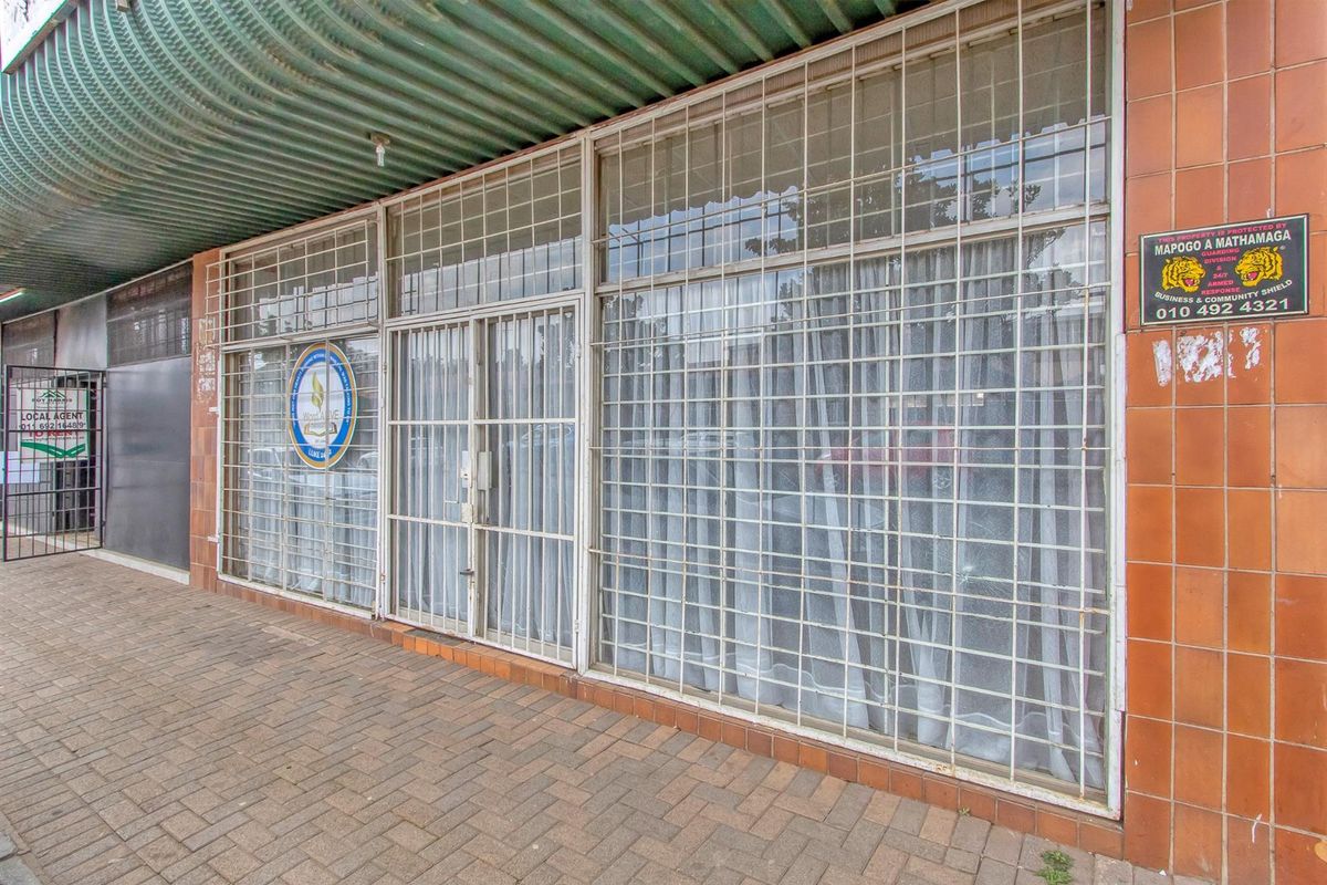 Commercial storefront featuring a tiled facade, large glass windows with security bars, and a corrugated metal awning over a paved walkway.