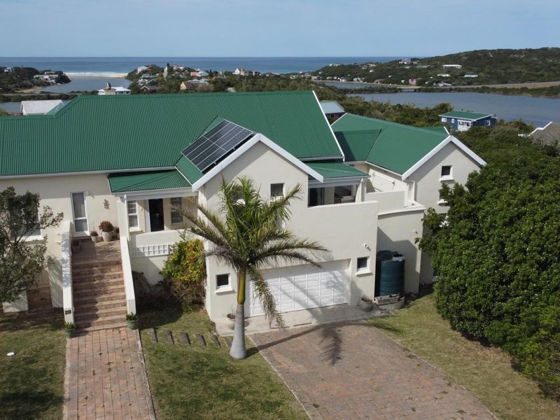 Aerial view of a coastal residence featuring a green metal roof, solar panels, paved driveway, and double garage, set in a scenic landscape.