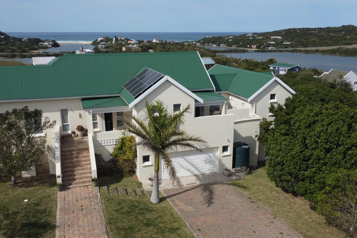 Aerial view of a coastal residence featuring a green metal roof, solar panels, paved driveway, and double garage, set in a scenic landscape.