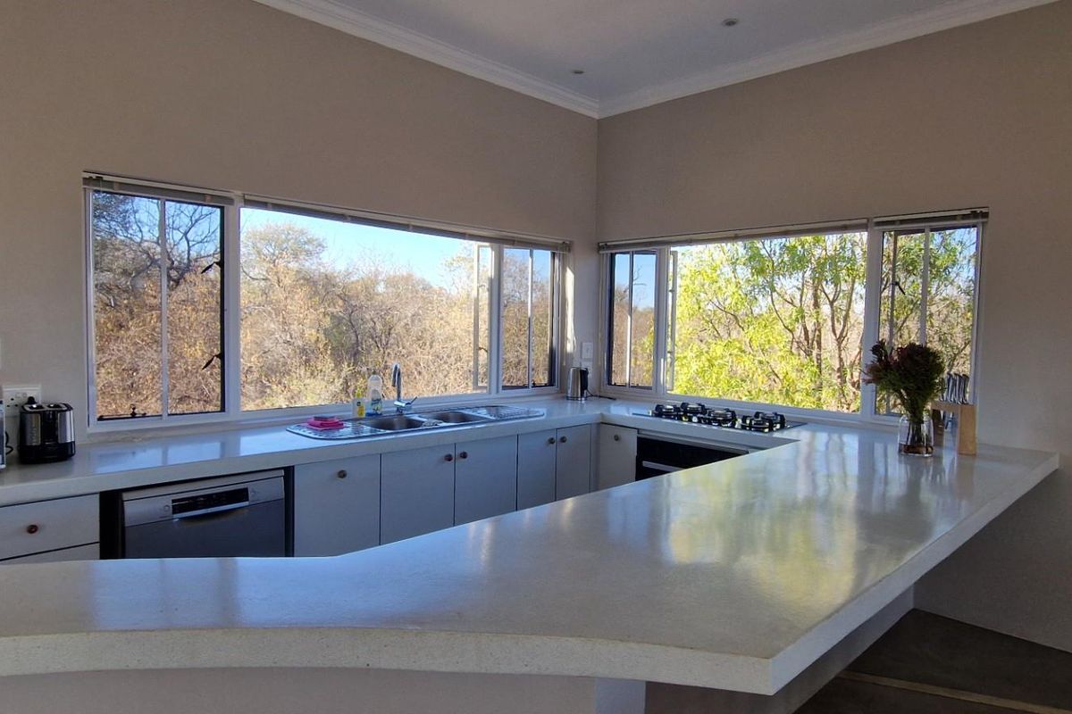 A bright kitchen featuring a wrap-around stone countertop, integrated gas hob, double sink, and large windows offering scenic views of the surrounding landscape.