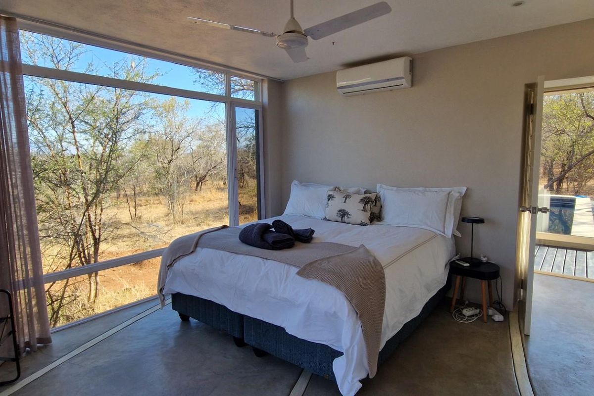 A bright bedroom featuring large floor-to-ceiling windows with bushveld views, polished concrete flooring, built-in cupboards, and a wall-mounted air conditioning unit.