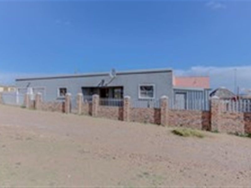 Front exterior view of a single-story house featuring a brick boundary wall with metal fencing and an unpaved approach.