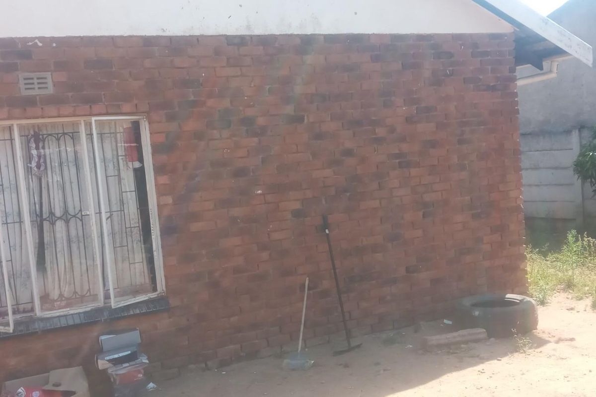 Exterior view of a brick-walled house featuring a window with security bars and a surrounding unpaved yard area.