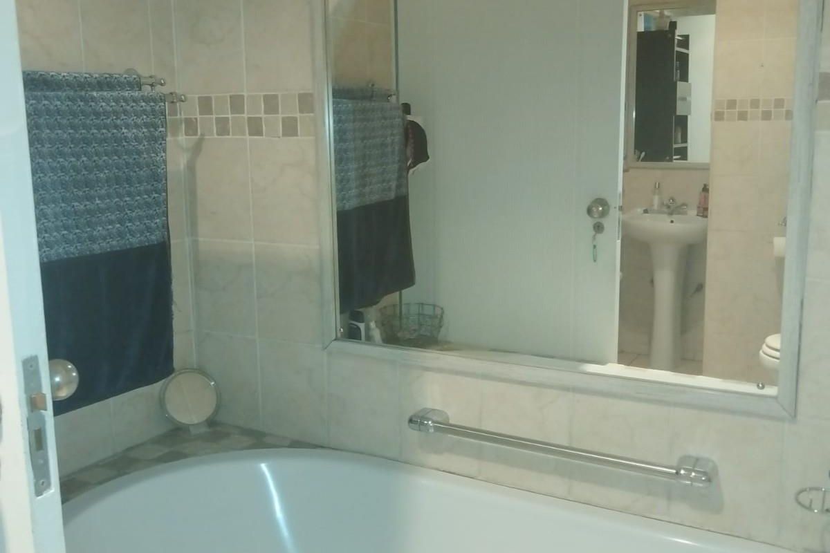 A functional bathroom featuring a built-in bathtub, wall-mounted grab rail, large vanity mirror, and pedestal basin with neutral wall tiling.