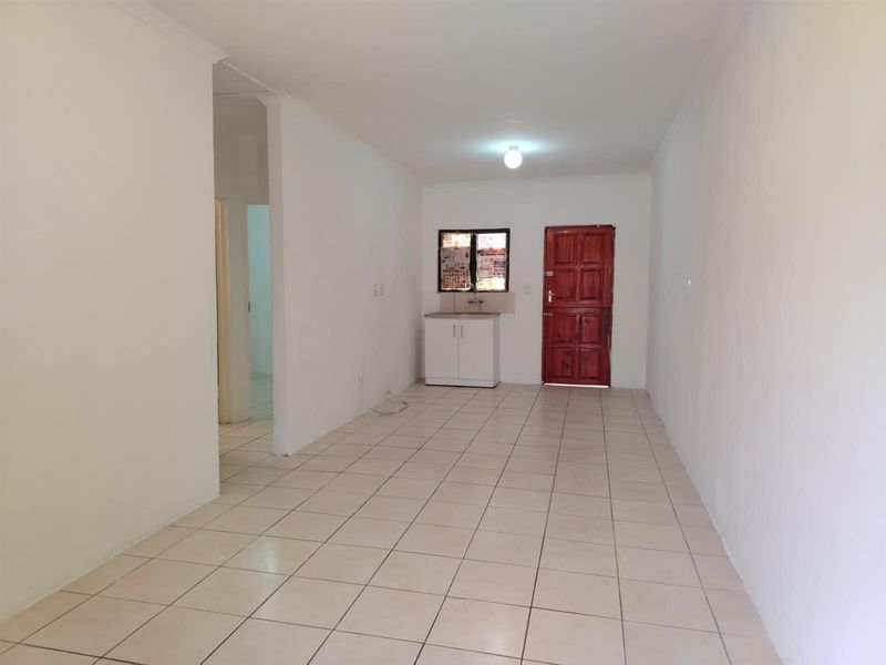 An empty, open-plan living area featuring tiled flooring, a small kitchenette unit, and a solid timber entrance door.
