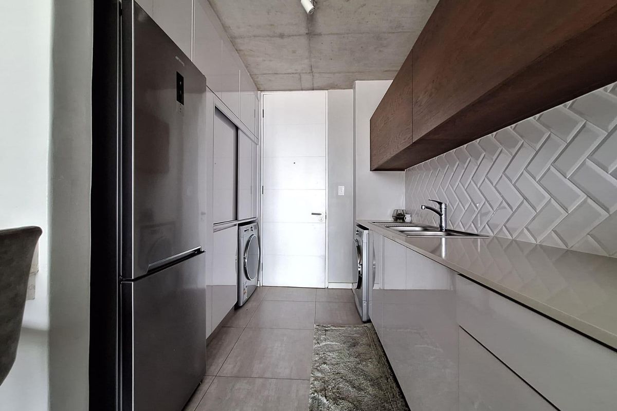Modern galley-style kitchen featuring high-gloss cabinetry, stone countertops, herringbone tile backsplash, and exposed concrete ceiling with integrated laundry appliances.