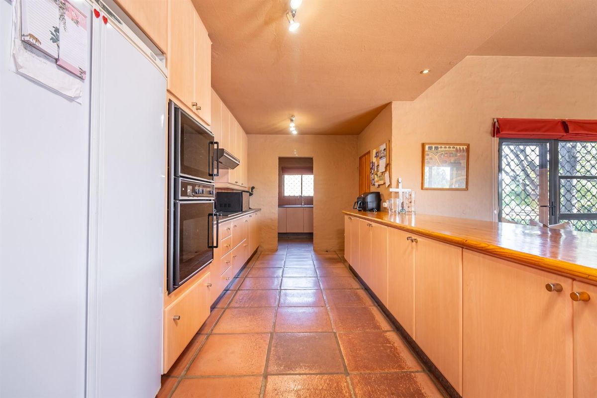 A functional kitchen featuring light wood cabinetry, built-in double ovens, and terracotta floor tiles, with a breakfast bar and direct access to a scullery area.