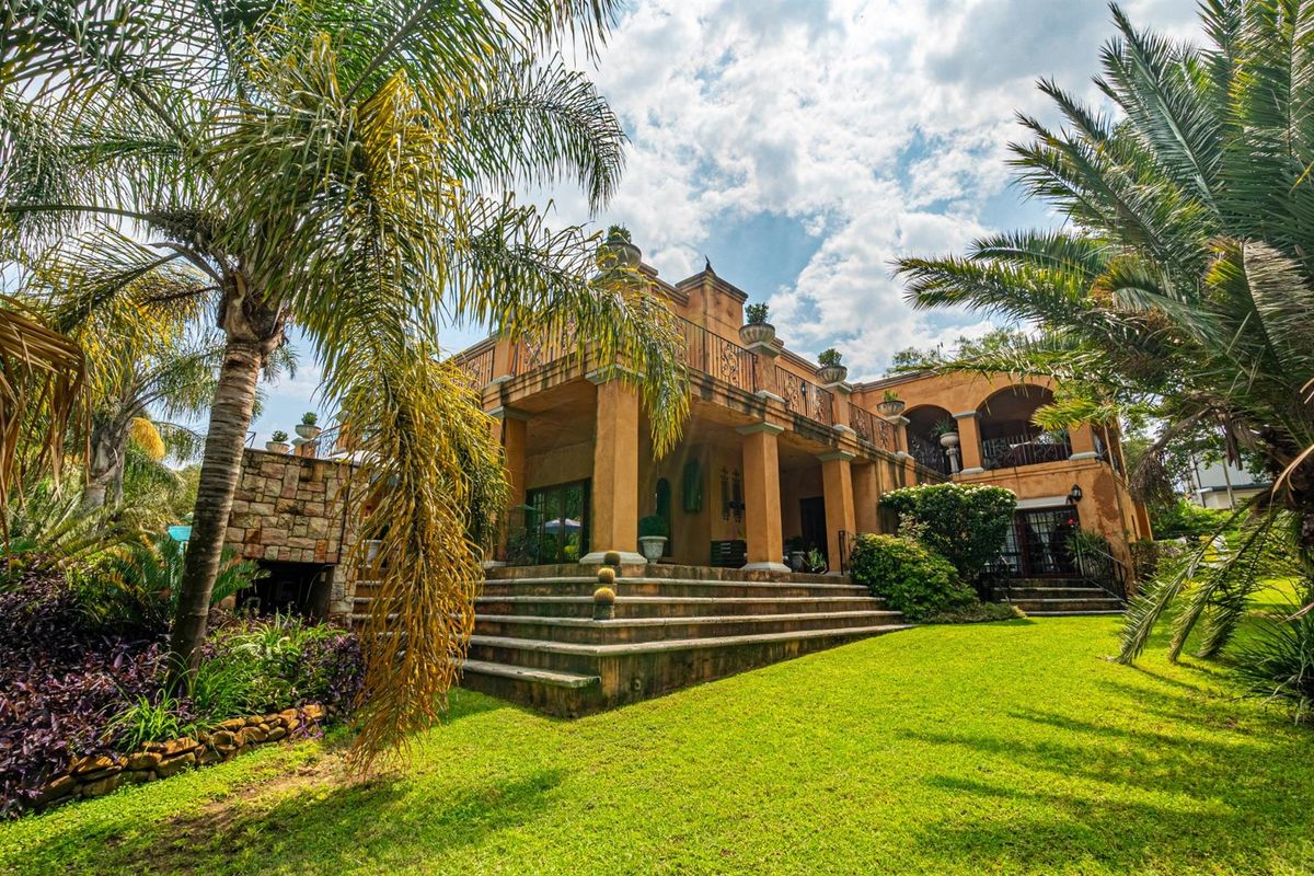 A Mediterranean-style rear elevation featuring a spacious covered patio, stone staircase, and established garden landscaping with mature palm trees.