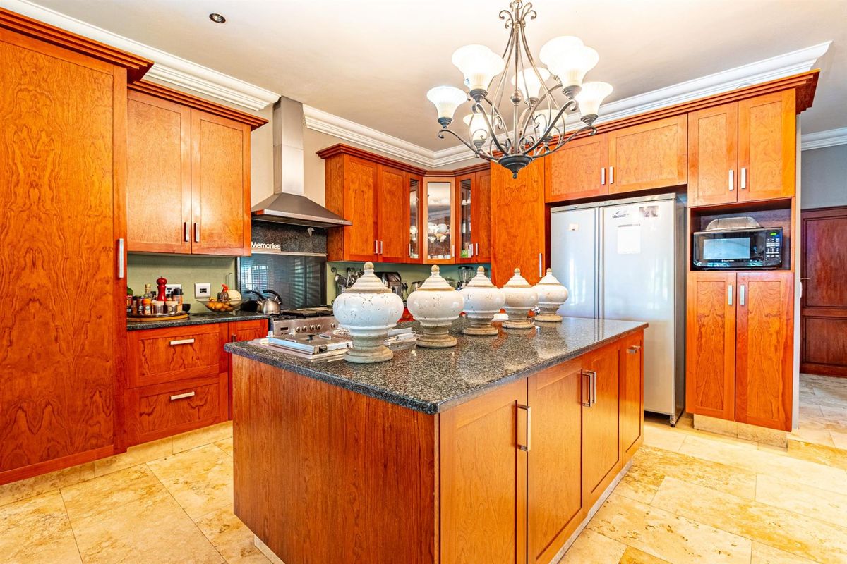 Spacious kitchen featuring warm-toned wooden cabinetry, a large central island with granite countertops, stainless steel extractor fan, and tiled flooring.