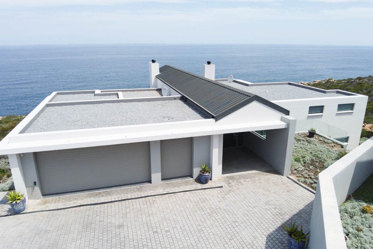 Aerial view of a modern coastal residence featuring a double garage, paved driveway, and a pitched metal roof overlooking the ocean.
