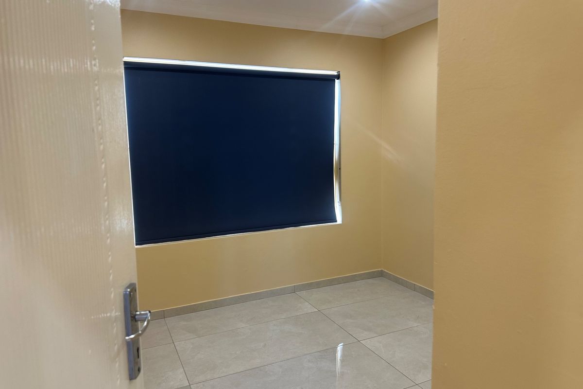 A clean, empty bedroom featuring neutral wall paint, tiled flooring, recessed ceiling lighting, and a fitted roller blind.