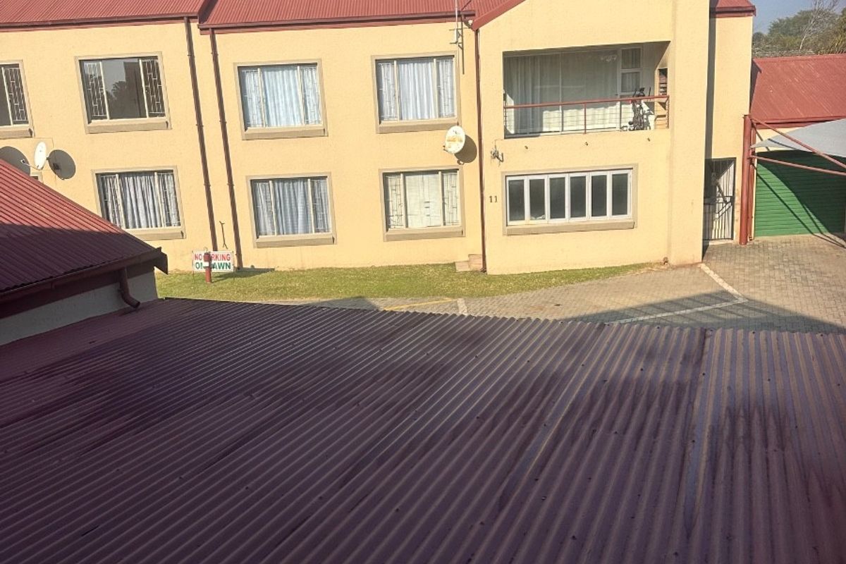 An elevated view of an apartment complex exterior featuring a corrugated metal roof, paved driveway, and a balcony on the upper level.