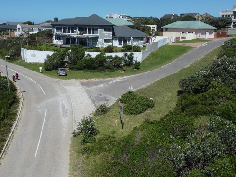 An aerial view of a vacant residential plot situated at a street corner, featuring natural vegetation and proximity to established neighborhood housing.
