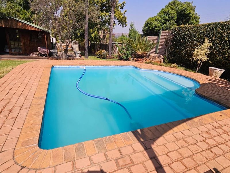 A well-maintained swimming pool area featuring brick paving, a surrounding garden, and a wooden outbuilding in the background.