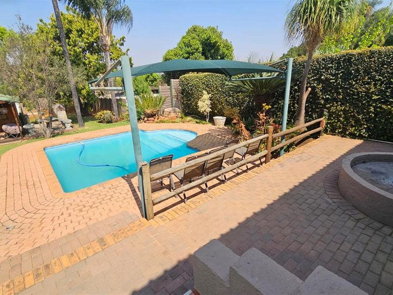 Outdoor pool area featuring a paved patio, built-in fire pit, shade sail, and established garden surroundings.