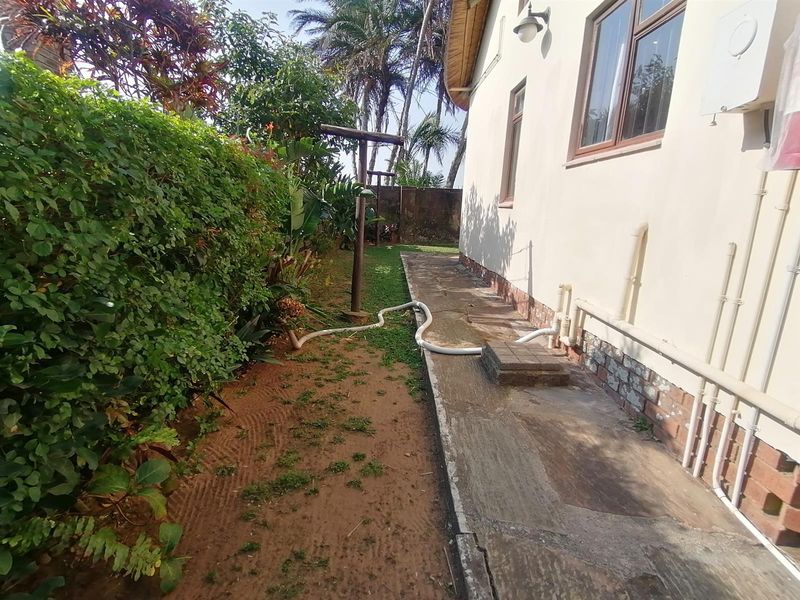 A side exterior view of the townhouse featuring a paved walkway, established garden hedge, and exposed external plumbing infrastructure.