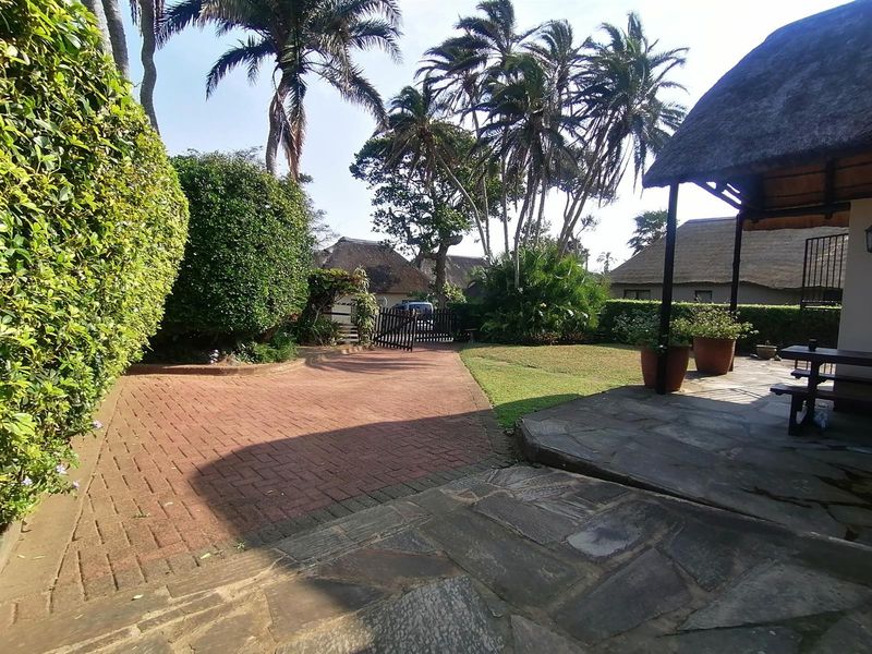 A spacious rear garden area featuring a stone-paved patio, a thatched roof outdoor structure, and a brick-paved driveway leading to a gated entrance.