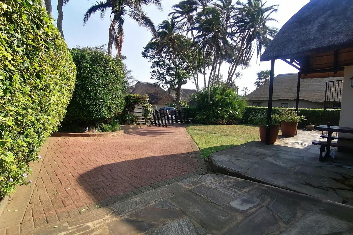 A spacious rear garden area featuring a stone-paved patio, a thatched roof outdoor structure, and a brick-paved driveway leading to a gated entrance.
