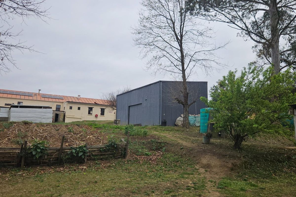 Exterior view of the small holding featuring a large grey metal storage shed, adjacent main building with solar panels, and surrounding garden area.