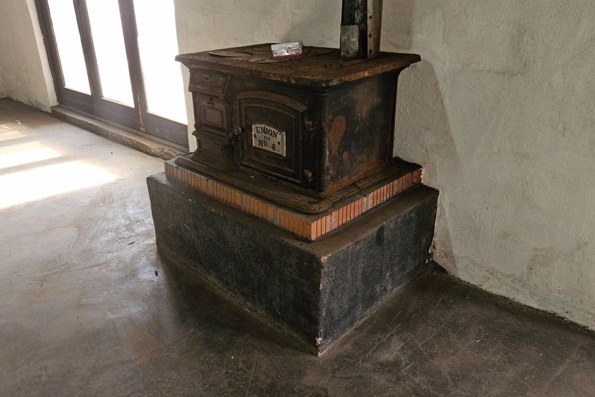 A rustic interior space featuring a vintage cast iron stove set on a raised masonry hearth with concrete flooring and natural light.