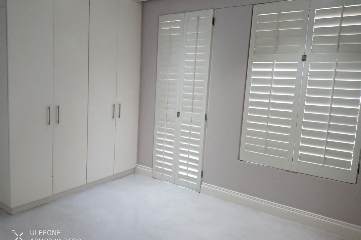 A bright bedroom featuring built-in floor-to-ceiling wardrobes and white plantation shutters on the windows, providing a clean and neutral living space.