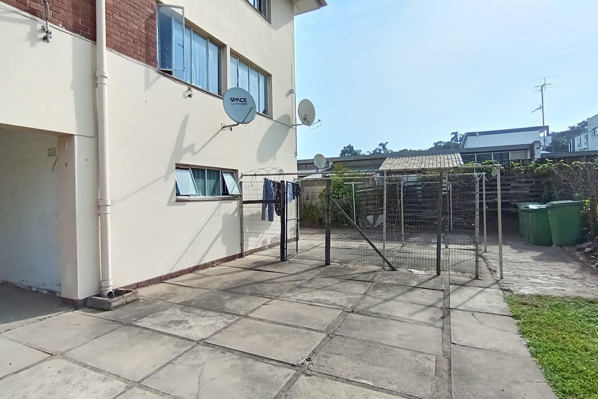 A rear exterior view of the apartment building featuring a paved courtyard area, metal security fencing, and a small covered utility structure.