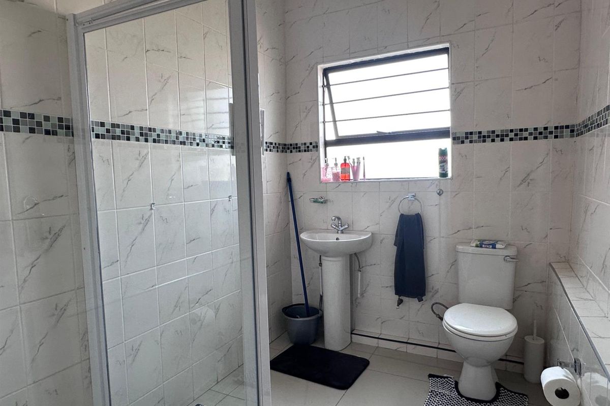 A functional bathroom featuring a glass-enclosed shower, pedestal basin, and toilet, finished with marble-look wall tiles and a decorative mosaic border.