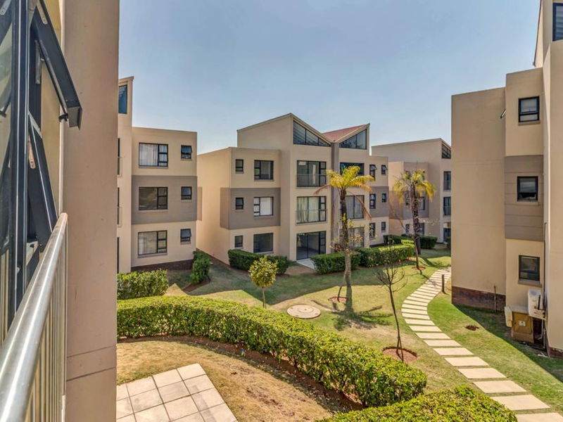 View of the communal garden area between apartment blocks, featuring paved walkways, manicured hedging, and established trees within the residential complex.