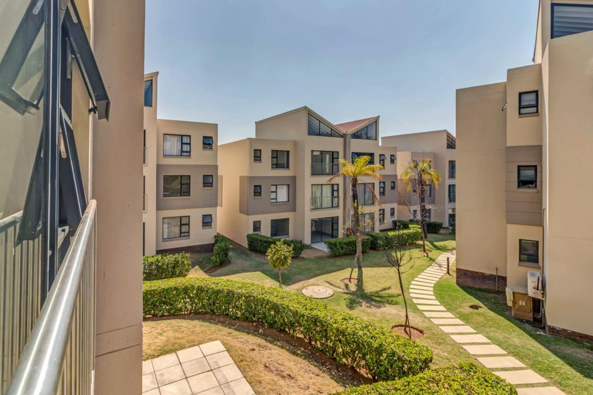 View of the communal garden area between apartment blocks, featuring paved walkways, manicured hedging, and established trees within the residential complex.